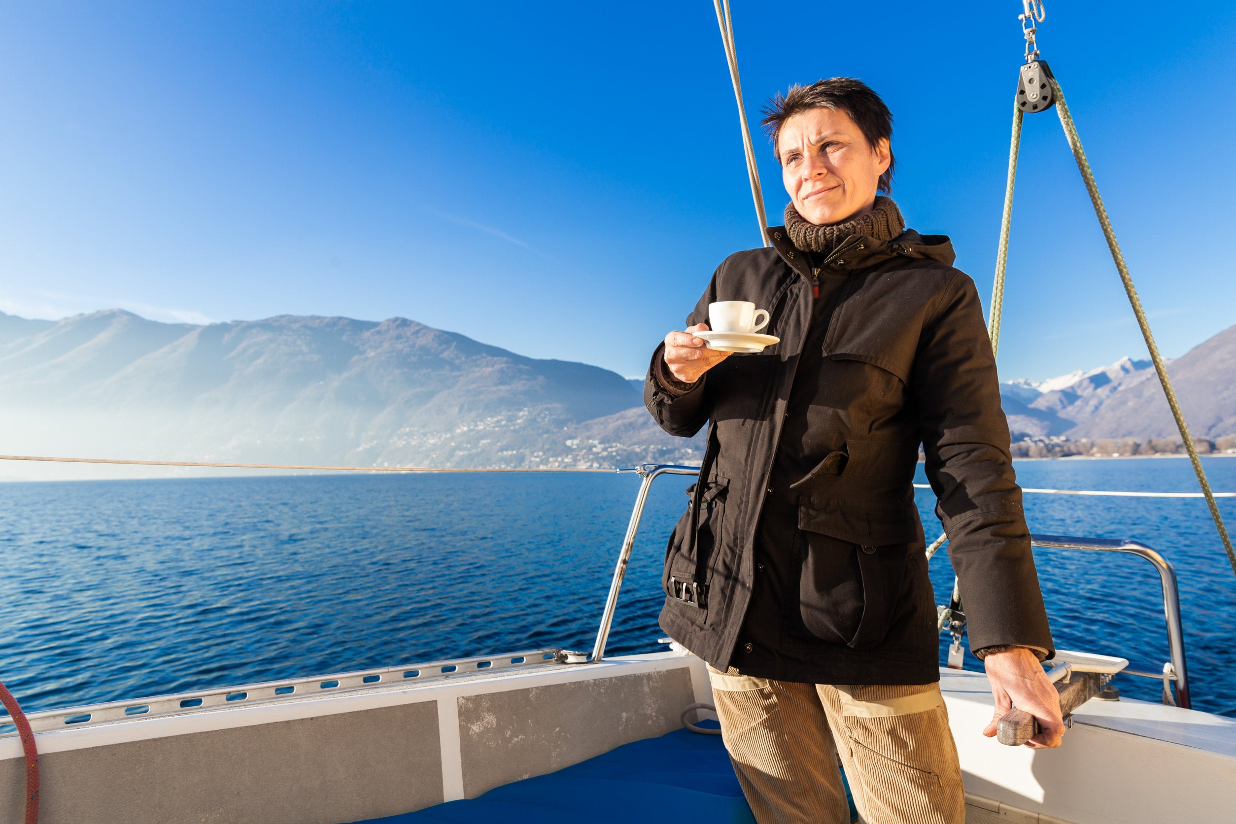 Confident-looking woman on a boat holding a coffee cup with mountains and blue sky in the background