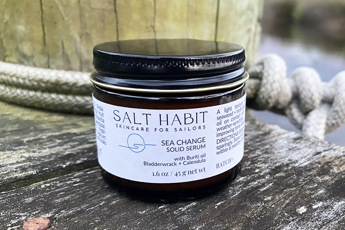 A jar of 'Salt Habit Skincare for Sailors Sea Change Pressed Serum' with an amber glass body and an aluminum lid, labeled with product information, placed on a wooden surface with a rope in the background.